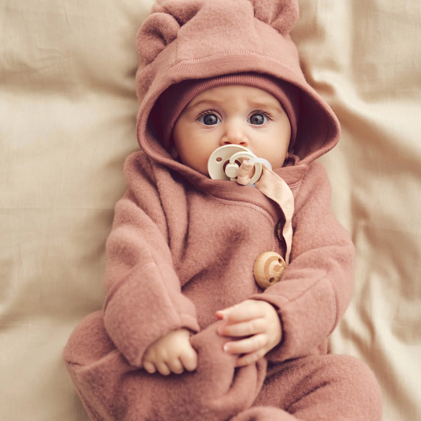 Baby wearing pink Wollwalk Merinowolle fleece suit with hood, lying on beige background.